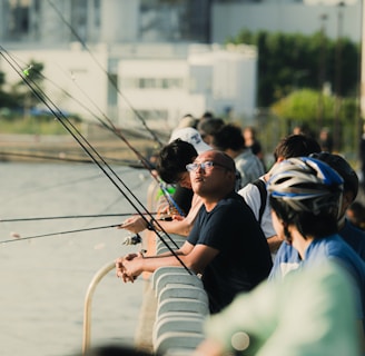 Several people are lined up along a railing by a body of water, engaged in fishing. Each person holds a fishing rod, with the lines cast into the water. The background shows an industrial or urban setting with buildings and greenery. The scene captures both leisure and concentration.